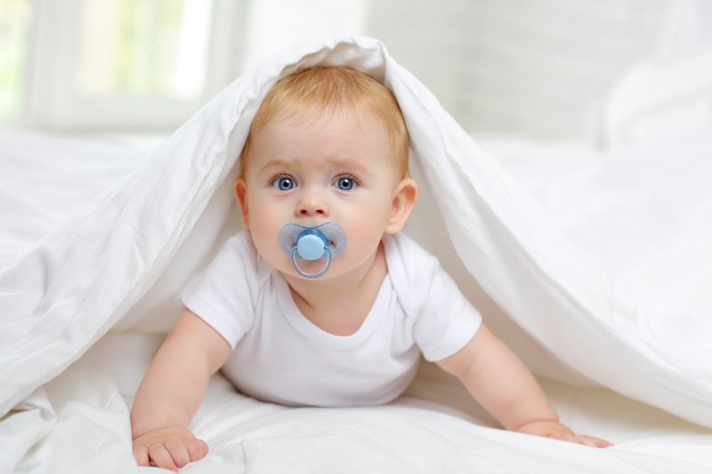 A baby sucking a pacifier beneath white sheets